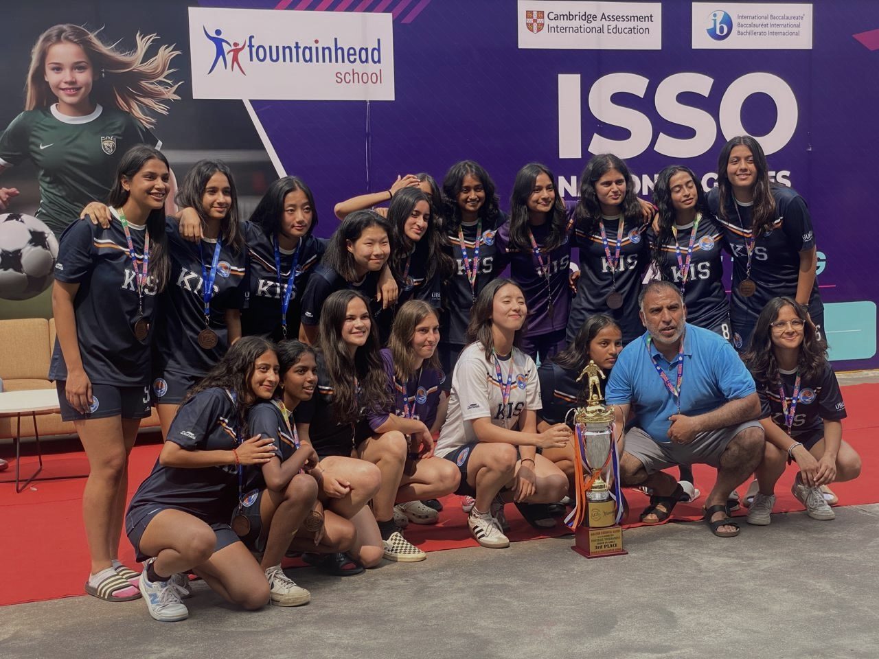 A girls' football team in navy kit with medals and a trophy at the ISSO National Games