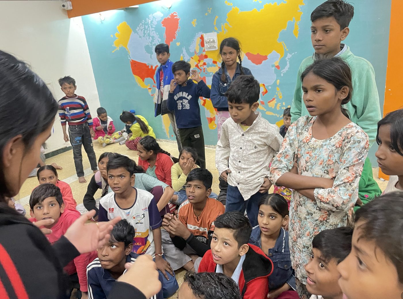Children listening closely to a facilitator in front of a world map mural